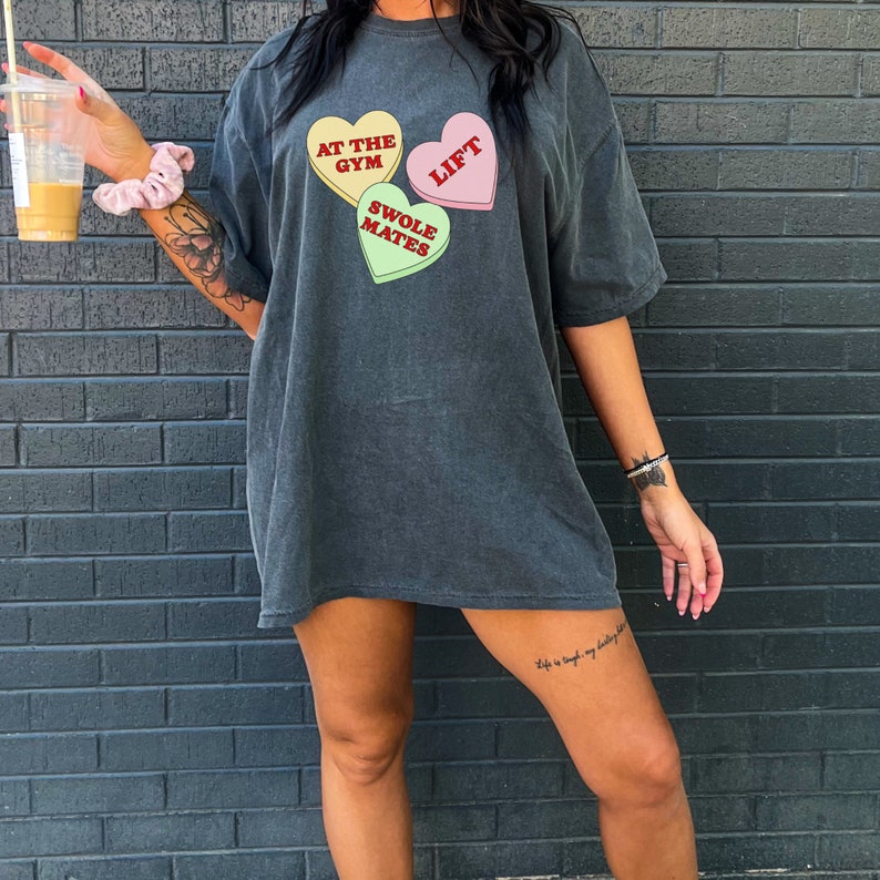 May include: A dark gray oversized t-shirt with a graphic of three candy hearts. The hearts say "At the Gym", "Swole Mates", and "Lift".