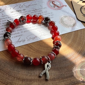 May include: Red and brown beaded bracelet with a silver charm in the shape of a ribbon. The bracelet is on a wooden surface with a card that says "Blood Clot Survivor".