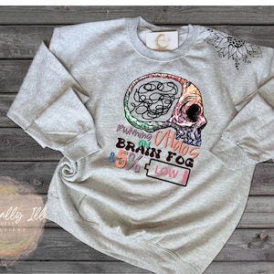 May include: Gray sweatshirt with a colorful graphic of a skull with a brain inside. The text "Running on Chaos, Brain Fog & 5% Low" is printed below the skull.