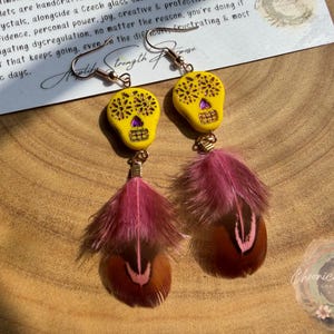 May include: Yellow skull earrings with purple accents and dangling feathers in shades of pink and brown. The earrings have a Day of the Dead theme. The earrings are hanging on a wooden surface.