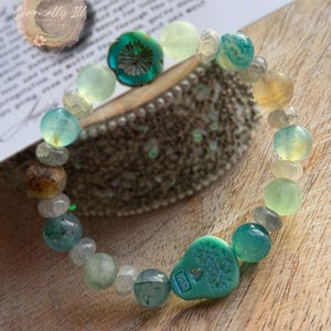 May include: A beaded bracelet featuring a variety of colorful gemstones in shades of green, blue, and yellow. The bracelet includes a turquoise-colored focal bead with a floral design and the text "Chronically Ill Goddess Designs".