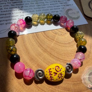 May include: A beaded bracelet featuring a mix of pink, black, and yellow beads. The bracelet includes a large yellow bead with a design and silver spiral accents. The background includes a wooden surface and a card with text.