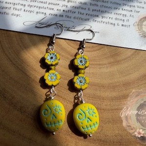 May include: A pair of handcrafted earrings featuring yellow and light blue floral beads and skull-shaped pendants. The earrings are made with silver-colored metal hooks and are displayed on a wooden surface.