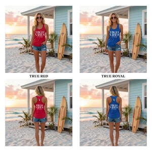 May include: A woman wearing a red tank top with the text "HIGH VIBES" in white. She is standing on a sandy beach in front of a light blue building and a surfboard. The tank top is paired with denim shorts.