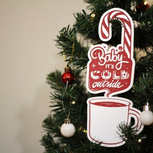 Può includere: Addobbo per albero di Natale rosso e bianco a forma di bastoncino di zucchero con una tazza di cioccolata calda e il testo "Baby it's cold outside".