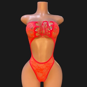 May include: A neon orange lace bodysuit featuring a strappy lace-up front detail, a deep cutout at the midsection, and scattered clear rhinestones throughout the fabric. The garment has a high-cut leg design and a sleeveless, strapless silhouette.