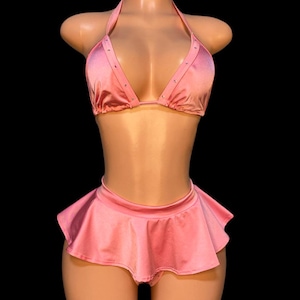 May include: A pink satin lingerie set on a mannequin. The set includes a halter-top bikini with rhinestone accents and a ruffled mini skirt. The fabric has a slight sheen, and the set is displayed against a black background.