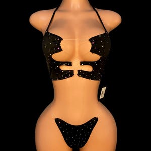 Op de afbeelding: Zwarte tweedelige outfit met een haltertop en bikinibroekje. De top heeft uitgesneden details en de set is versierd met kleine, sprankelende accenten. Doorzichtige bandjes verbinden de top en de broek.