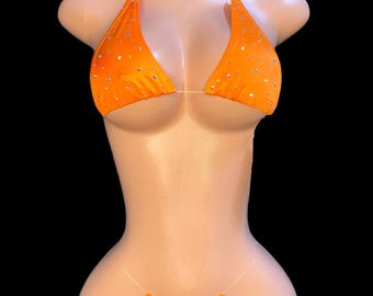 Puede incluir: Conjunto de bikini naranja brillante con pedrería. El top triangular y la braguita de tanga se muestran en un maniquí sobre un fondo negro. El bikini está hecho de un tejido brillante.