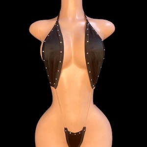 Puede incluir: Bikini negro con cuello halter y detalles de pedrería. La parte superior e inferior del bikini están conectadas por finas correas. El bikini se exhibe en un maniquí sobre un fondo negro.
