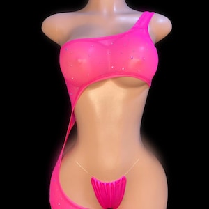 May include: A bright pink, one-shoulder bodysuit with a sheer mesh overlay and sparkling embellishments. The bodysuit features a cutout design and a matching thong bottom, displayed on a mannequin against a black background.