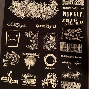 May include: Black fabric with white graphic designs and text. The text includes band names like "orchid", "foxtails", and "NOVELY.", along with other phrases and illustrations. The overall design has a punk rock aesthetic.