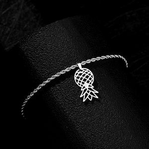 May include: A silver-toned anklet featuring a pineapple pendant. The pendant has a lattice design for the fruit and detailed leaves. The anklet chain has a twisted rope design. The jewelry is displayed against a dark background.