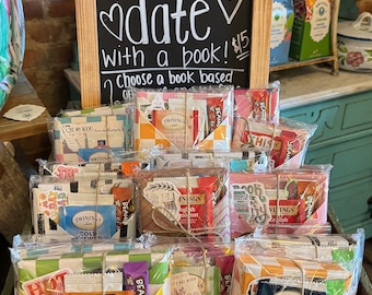 May include: A wooden crate filled with wrapped gifts, each containing a book and a box of tea. The gifts are tied with twine and decorated with colorful paper. A chalkboard sign above the crate reads "Blind date with a book! $15 Choose a book based on..."