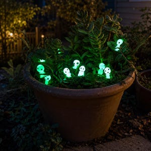 May include: A terracotta-coloured planter filled with green succulent plants and several small, glowing white figurines with round heads and simple facial features. The figurines emit a bright green light, creating a luminous effect among the leaves.