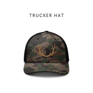 May include: A black and green camouflage trucker hat with a gold embroidered antler design. "TRUCKER HAT" is printed in black text above the hat.