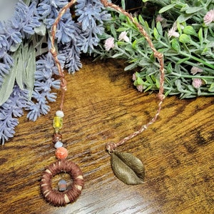 May include: A brown and beige cord necklace with a small, round, woven pendant, a leaf charm, and colorful beads.