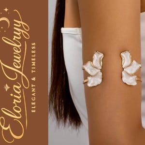 May include: Gold-toned arm cuff with white floral accents. The jewelry features a delicate design with a glossy finish. The brand name "Eloria Jewelryy" is displayed in elegant script, with the tagline "Elegant & Timeless".