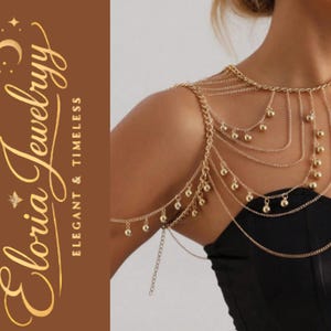 Puede incluir: Cadena de cuerpo dorada con múltiples hilos y pequeñas esferas colgantes. La joya está diseñada para cubrir los hombros y el brazo, creando un efecto de capas. El texto "Eloria Jewelryy Elegant & Timeless" es visible.