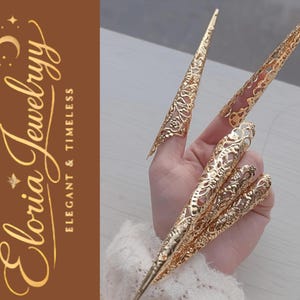 Puede incluir: Cubiertas de uñas doradas y ornamentadas con intrincados diseños de filigrana. Las cubiertas de uñas son largas y puntiagudas, creando un efecto dramático. La imagen también presenta el texto "Eloria Jewelryy Elegant & Timeless."