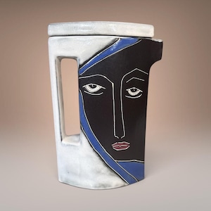 May include: A ceramic pitcher featuring a stylized face design. The vessel has a white handle and base, a black face with white line details, a blue accent stripe, and red lips. The pitcher has a flat top and a small spout on the right side.