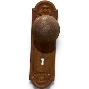 May include: A weathered, rust-colored metal door knob set featuring a spherical knob attached to a rectangular backplate. The backplate displays decorative embossed floral motifs at the top and bottom ends and includes a central keyhole opening.