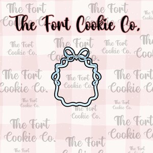 May include: A light blue cookie cutter in the shape of a bib with a bow at the top. The text "The Fort Cookie Co." is displayed in a cursive font.