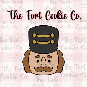 May include: A cookie cutter in the shape of a nutcracker's face, featuring a black hat with gold accents, a brown face, and a mustache. The text "The Fort Cookie Co." is displayed in a cursive font.