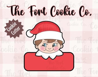 May include: A red and white cookie cutter shaped like an elf with a Santa hat. The cutter has a blank space for adding a name. The text "The Fort Cookie Co." is at the top of the image.
