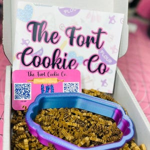 May include: A box from "The Fort Cookie Co." contains a blue and purple cookie cutter. The box also contains a card with the company's name and logo, and is filled with brown crinkle paper.