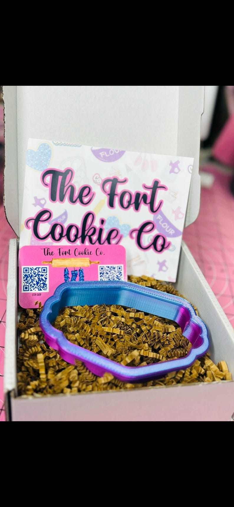 May include: A box from "The Fort Cookie Co." contains a blue and purple cookie cutter. The box also contains a card with the company's name and logo, and is filled with brown crinkle paper.