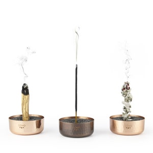 May include: Three copper incense holders with different types of incense burning. The holders are hammered copper and have the word "COHO" engraved on the front.