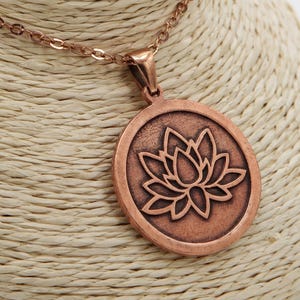May include: Copper-colored necklace featuring a round pendant with a lotus flower design. The pendant is encircled by a raised border and hangs from a delicate chain. The necklace is displayed on a beige bust.