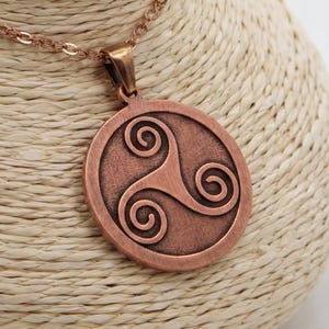 May include: A copper-toned pendant necklace with a circular pendant displaying a Celtic triskele design. The necklace is presented on a mannequin bust. The chain and pendant share a metallic finish, creating a coordinated and fashionable accessory.