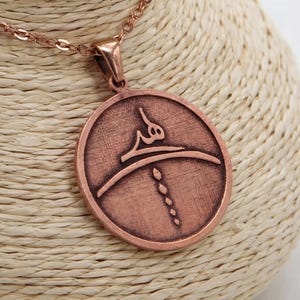 Op de afbeelding: Een koperkleurige hangerketting. De hanger heeft een cirkelvorm met een reli&euml;f ontwerp. De ketting en hanger hebben een bijpassende koperkleur. De ketting wordt gepresenteerd op een beige buste.