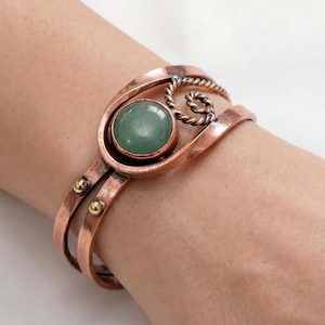May include: Copper cuff bracelet featuring a large, round, pale green stone set in an openwork design. The bracelet has a double band and is adorned with small gold-colored rivets and a twisted metal accent.