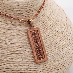 Puede incluir: Un collar de tono cobre con un colgante rectangular que presenta un patr&oacute;n de enredadera floral en relieve. El colgante cuelga de una delicada cadena a juego, mostrando un acabado met&aacute;lico y un dise&ntilde;o vertical con flores.