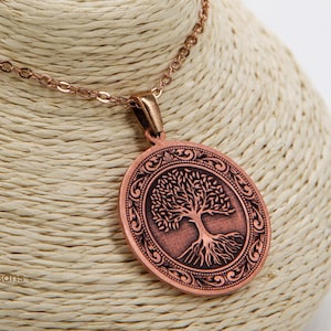 May include: A copper-colored necklace with a round pendant featuring a detailed tree design. The pendant is suspended from a delicate chain. The necklace is displayed on a beige bust. The text "Cu29 Series Handcrafted by Our Local Artisans" is visible.