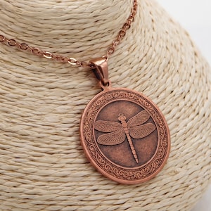 Pode incluir: Um colar com pendente circular em tom de cobre com uma grava&ccedil;&atilde;o detalhada de uma lib&eacute;lula no centro, rodeada por um padr&atilde;o de borda intrincado. O pendente est&aacute; suspenso numa corrente de metal a condizer com elos ovais uniformes.