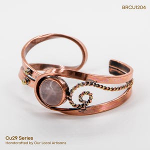 May include: Copper cuff bracelet with a light pink gemstone center. The bracelet features a twisted rope design and small gold-colored accents. The text "BRCU1204" is visible. The text "Cu29 Series Handcrafted by Our Local Artisans" is also visible.
