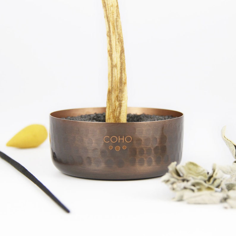 May include: A hammered copper incense holder with a wooden stick standing in the centre. The holder has the word "COHO" and three dots engraved on the side.