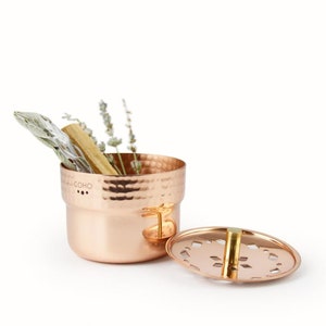 May include: A hammered copper incense burner with a lid featuring a decorative cut-out design. The burner is filled with dried herbs and a stick of palo santo wood. The text "COHO" is engraved on the side of the burner.