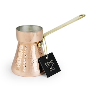 Puede incluir: Cafetera de color cobre con textura martillada y asa dorada. La cafetera tiene una parte superior cilíndrica y un cuerpo cónico. Una etiqueta negra con texto blanco cuelga del asa. La etiqueta dice "COHO objet originale."