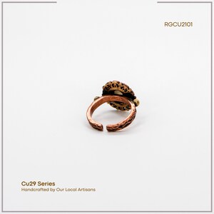 Puede incluir: Un anillo de color cobre con un dise&ntilde;o ornamentado de inspiraci&oacute;n floral. El anillo presenta una piedra central rodeada por un engaste de filigrana detallado. La banda tiene un patr&oacute;n texturizado. El texto "Cu29 Series" y "Handcrafted by Our Local Artisans" es visible.