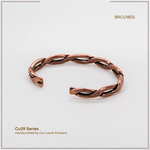 K&ouml;nnte beinhalten: Ein kupferfarbenes, offenes Armband mit geflochtenem Design. Das Armband besteht aus ineinander verschlungenen Metallstr&auml;ngen. Der Text "BRCU1805" und "Cu29 Series Handcrafted by Our Local Artisans" ist zu sehen.