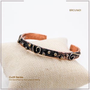 May include: Copper cuff bracelet with a dark, textured band, decorated with gold-coloured accents and swirling designs. The bracelet is part of the Cu29 Series, handcrafted by local artisans. It rests on a beige cushion.