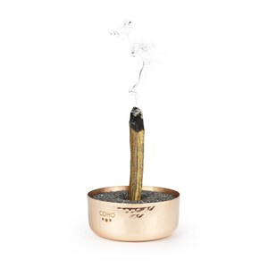 May include: A copper incense holder with a hammered finish and the word "COHO" engraved on the side. A wooden incense stick is burning in the holder, with smoke rising from the top.