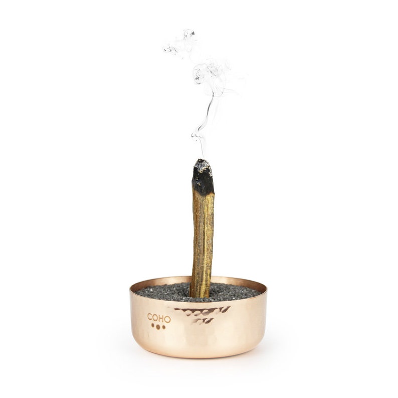 May include: A copper incense holder with a hammered finish and the word "COHO" engraved on the side. A wooden incense stick is burning in the holder, with smoke rising from the top.