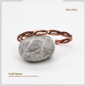 K&ouml;nnte beinhalten: Ein kupferfarbenes, geflochtenes Armband liegt auf einem grauen Stein. Das Armband hat ein verdrehtes Design und ist an einem Ende offen. Das Bild enth&auml;lt auch den Text "BRCU1805" und "Cu29 Series Handcrafted by Our Local Artisans."