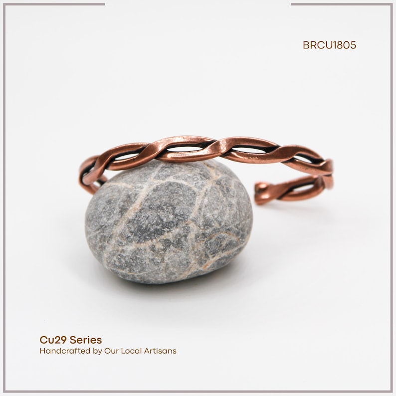 K&ouml;nnte beinhalten: Ein kupferfarbenes, geflochtenes Armband liegt auf einem grauen Stein. Das Armband hat ein verdrehtes Design und ist an einem Ende offen. Das Bild enth&auml;lt auch den Text "BRCU1805" und "Cu29 Series Handcrafted by Our Local Artisans."